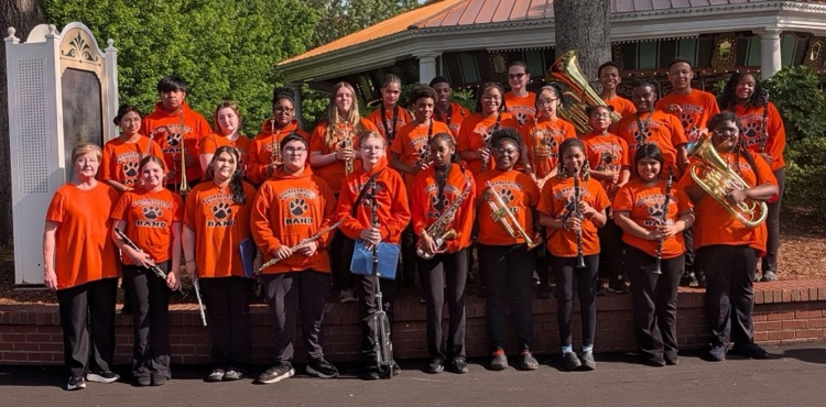 A group of students wearing matching orange shirts that say Elizabethtown Middle School Band, and holding various instruments stand together smiling.