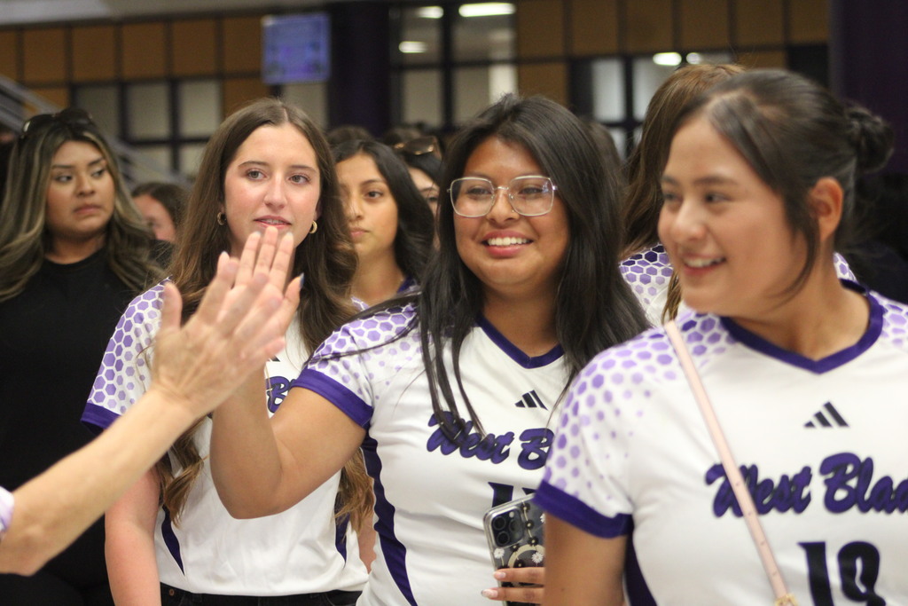 West Bladen Lady Knights varsity soccer players smile and greet supporters during a school send-off before their game against Wake Prep. Several players in white and purple jerseys walk through the crowd while students and staff cheer them on.
