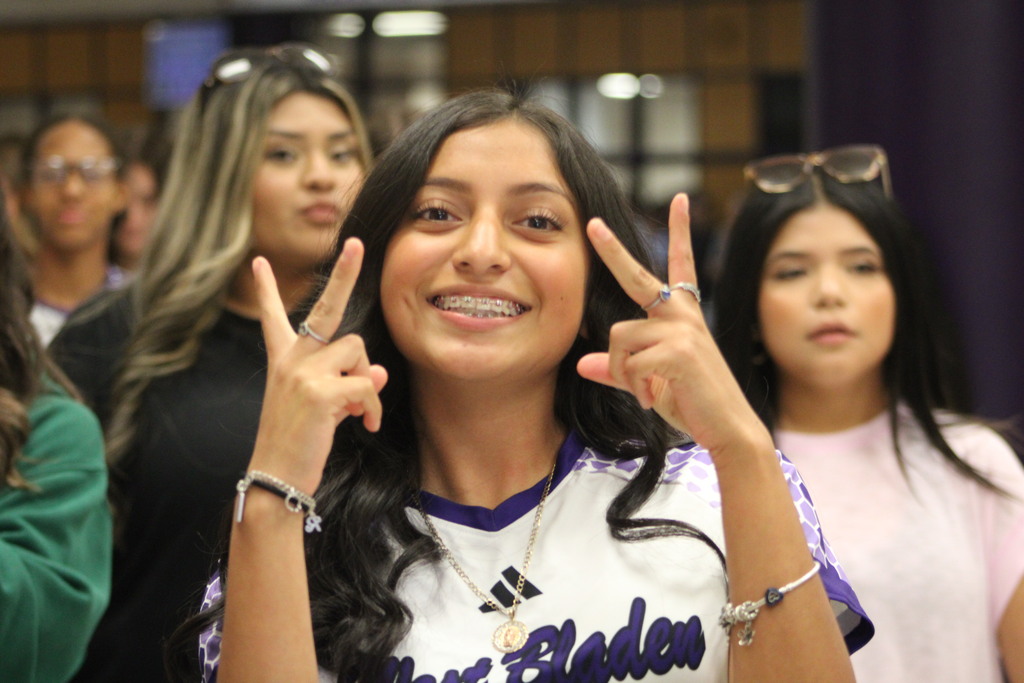 A smiling West Bladen Lady Knights varsity soccer player poses with peace signs during the team’s school send-off before traveling to face Wake Prep. Other students and teammates stand in the background of the school atrium.