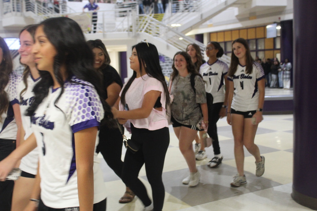 Members of the West Bladen Lady Knights varsity soccer team walk through the school commons during a team send-off before traveling to play Wake Prep. Students and staff line the atrium as the players smile and make their way through the building in their white and purple jerseys.