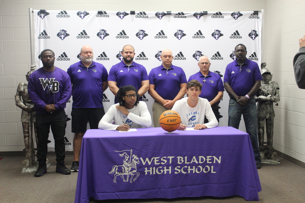 Jackson Pait and Tylik McCall sit at a table signing to continue their basketball careers at Chowan University while West Bladen coaches and staff stand behind them during the signing ceremony.