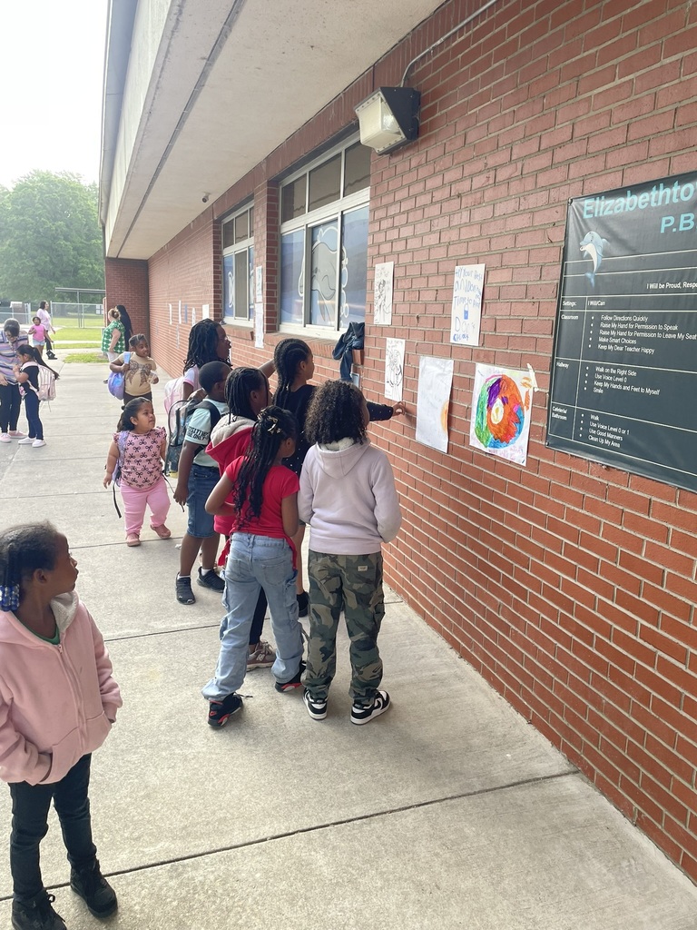 Students walk down a school sidewalk lined with handmade motivational posters and sidewalk displays encouraging 3rd and 4th graders before their Reading EOG. The brick wall is covered with colorful signs reminding students to try their best and stay confident.