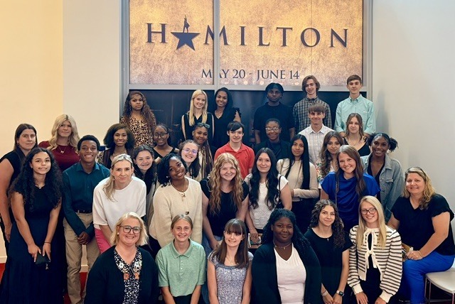 group photo taken in front of a poster for the musical Hamilton. The group includes a mix of teenagers and adults, likely students with teachers, chaperones, or program leaders. Everyone is posed indoors in several rows, and the atmosphere looks like a school trip, theater program, or educational outing connected to the show.