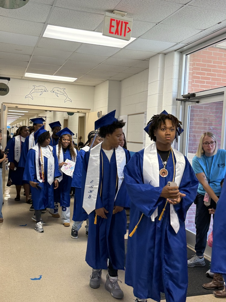 Graduating East Bladen seniors in blue caps and gowns walk through the halls of Elizabethtown Primary as younger students cheer and hold handmade Class of 2026 signs.