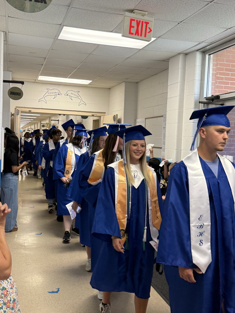 Graduating East Bladen seniors in blue caps and gowns walk through the halls of Elizabethtown Primary as younger students cheer and hold handmade Class of 2026 signs.