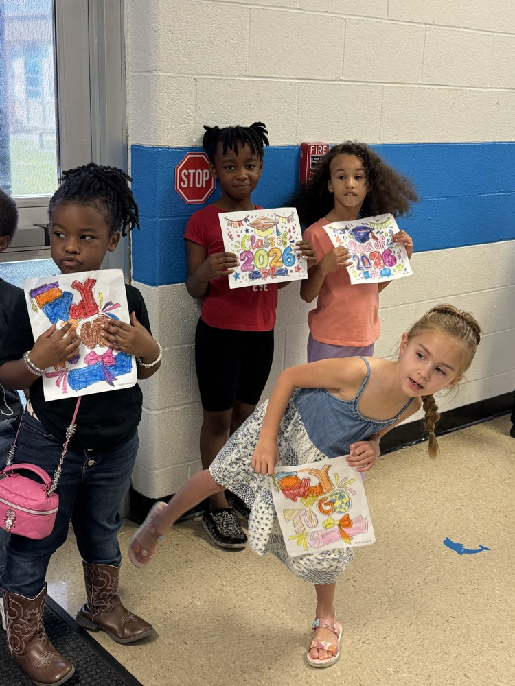 Graduating East Bladen seniors in blue caps and gowns walk through the halls of Elizabethtown Primary as younger students cheer and hold handmade Class of 2026 signs.