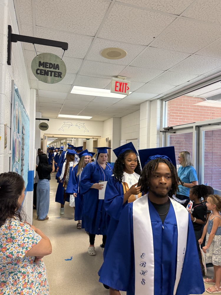 Graduating East Bladen seniors in blue caps and gowns walk through the halls of Elizabethtown Primary as younger students cheer and hold handmade Class of 2026 signs.