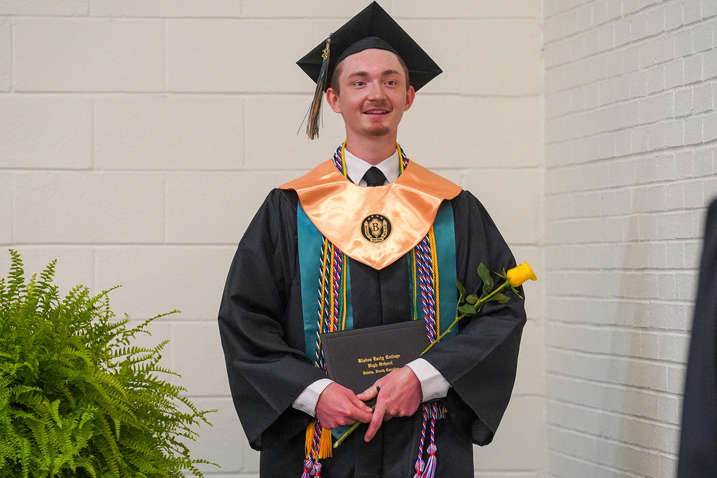 A smiling graduate in black cap and gown holds his Bladen Early College High School diploma and a yellow rose after receiving his degree.