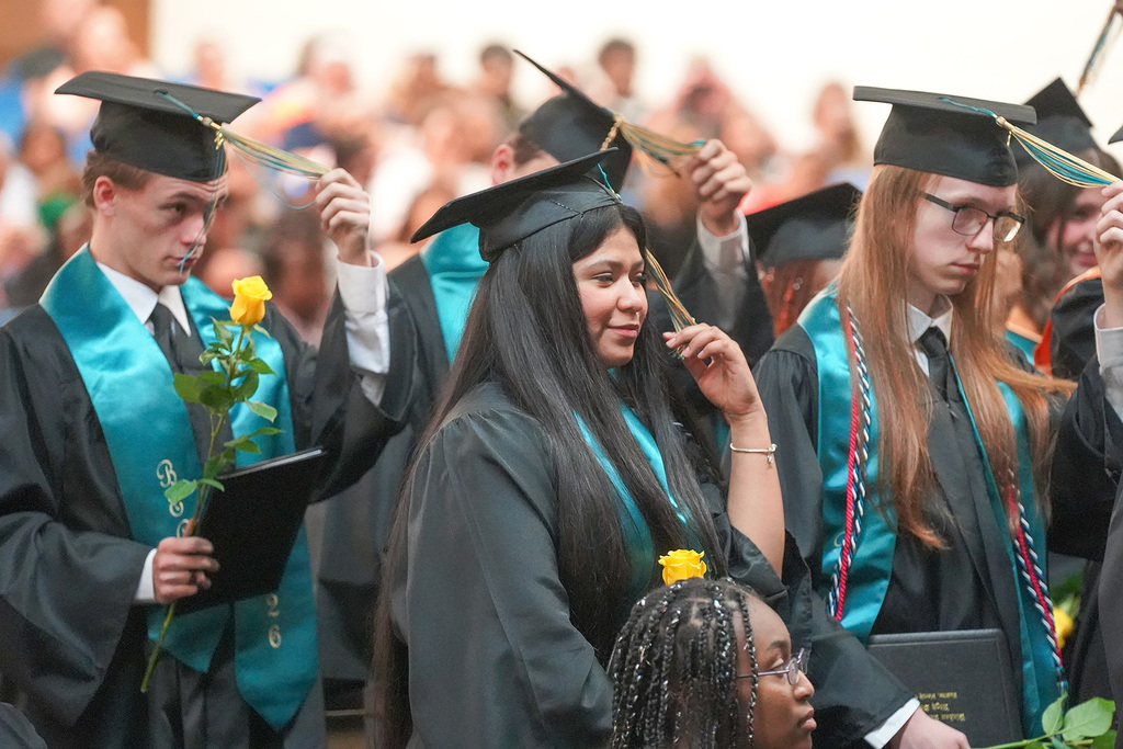 A group of graduates in black cap and gown move their tassels from right to left during the tassel-turning ceremony, holding yellow roses and diplomas.