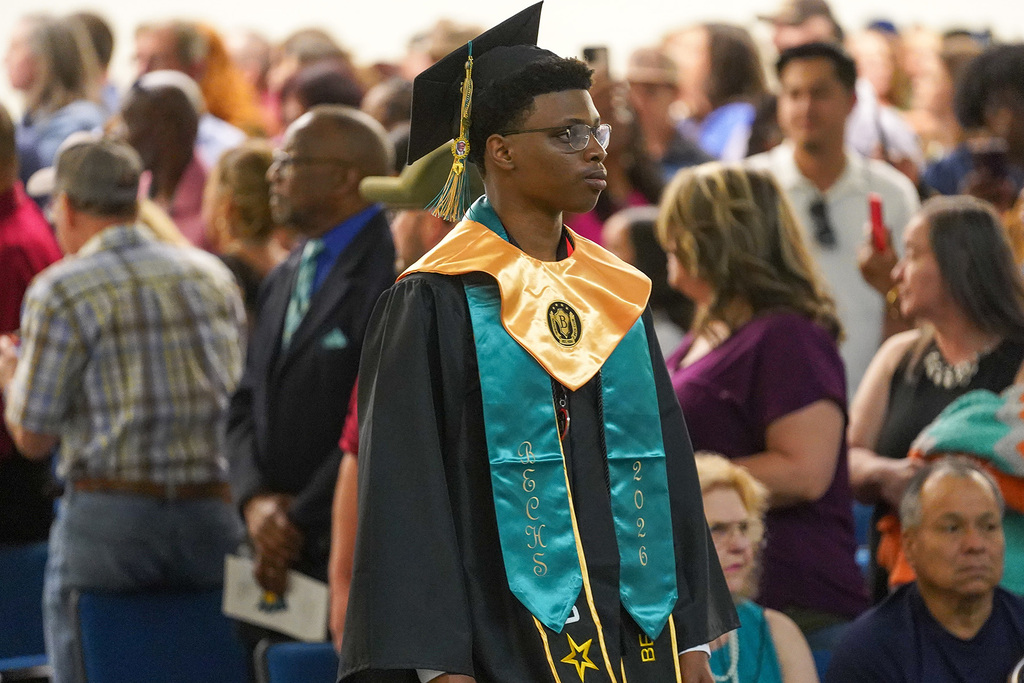 A graduate in black cap and gown with a teal BECHS 2026 stole and gold honor cord walks during the graduation processional as family members look on from the stands.