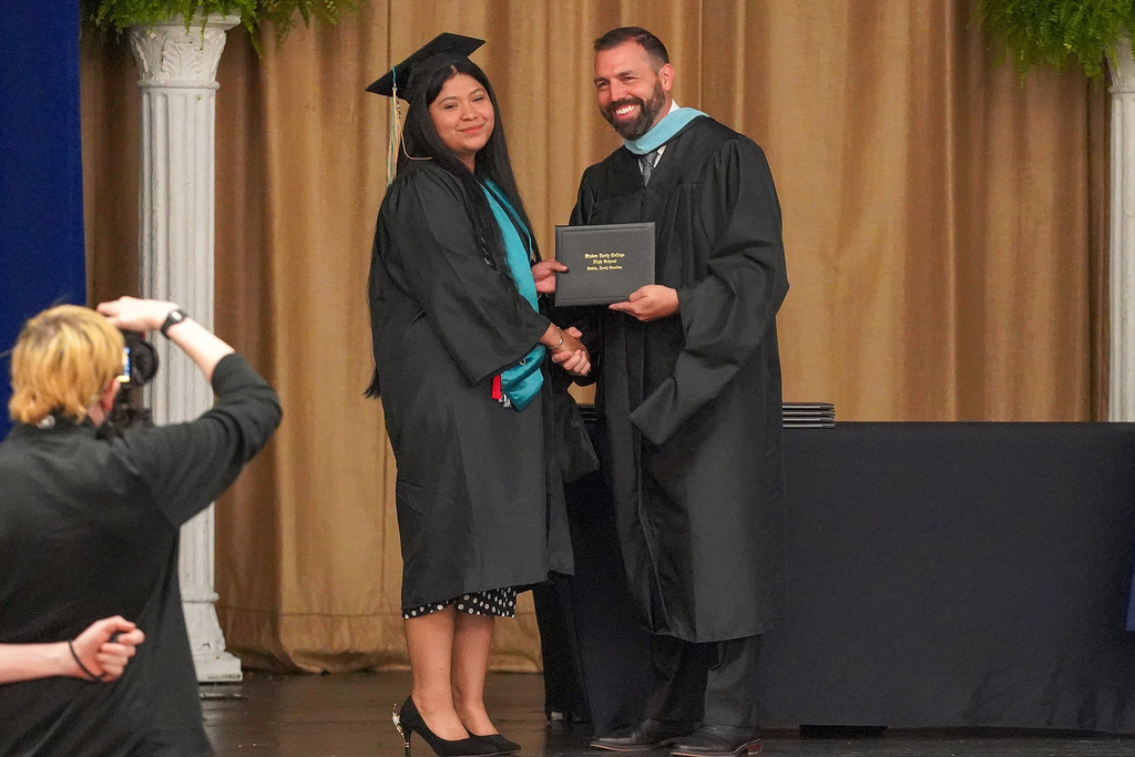 A graduate and a school official in academic regalia smile and pose together while jointly holding a Bladen Early College High School diploma on stage as a photographer captures the moment.