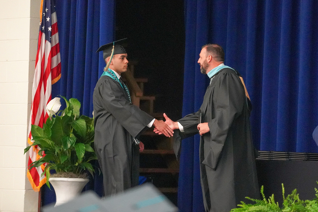 A graduate in black cap and gown shakes hands with a school official in academic regalia on stage during the diploma ceremony, with an American flag visible in the background.