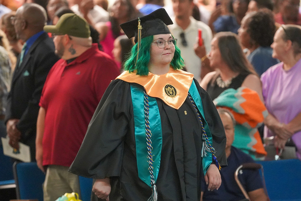 A graduate in black cap and gown with teal and gold regalia and green hair walks during a graduation processional through a crowded gymnasium.