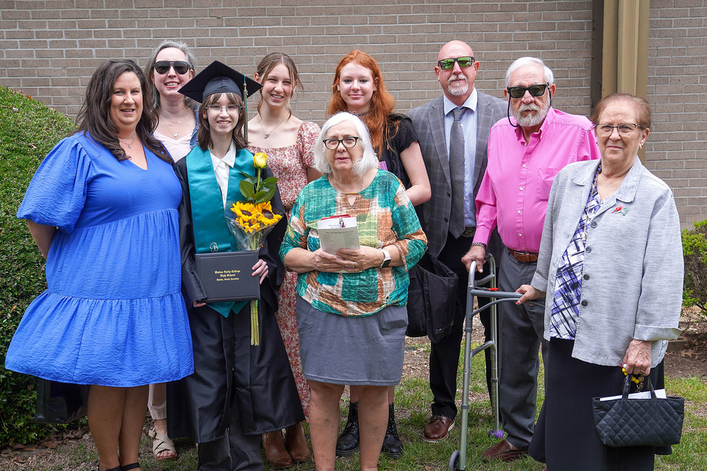 A graduate in black cap and gown holds sunflowers and a Bladen Early College High School diploma while posing outdoors with a large group of proud family members spanning multiple generations.