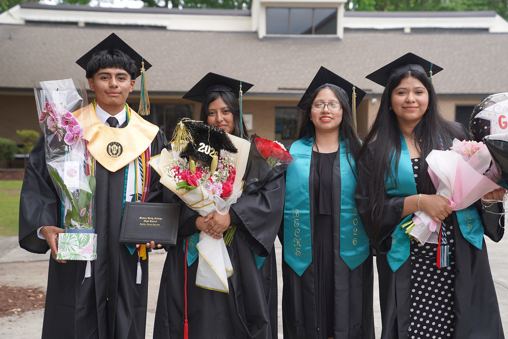 Four graduates in black cap and gown with BECHS 2026 teal stoles pose outdoors after the ceremony, holding bouquets of flowers and a diploma to celebrate their graduation day.
