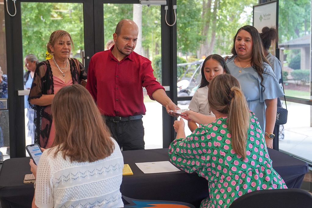 A family checks in at a registration table in a building lobby before a graduation ceremony. A man in a red shirt hands documents to a staff member in a green floral dress while family members look on.