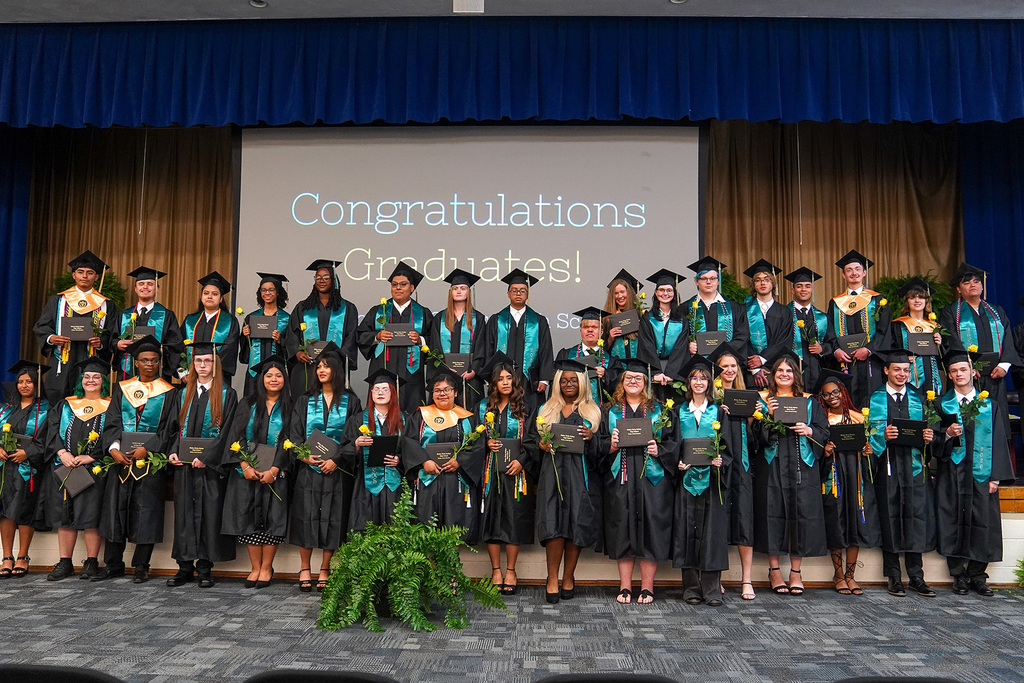 The full graduating class of Bladen Early College High School poses on stage in black cap and gown with teal stoles, holding diplomas and yellow roses in front of a "Congratulations Graduates!" screen.