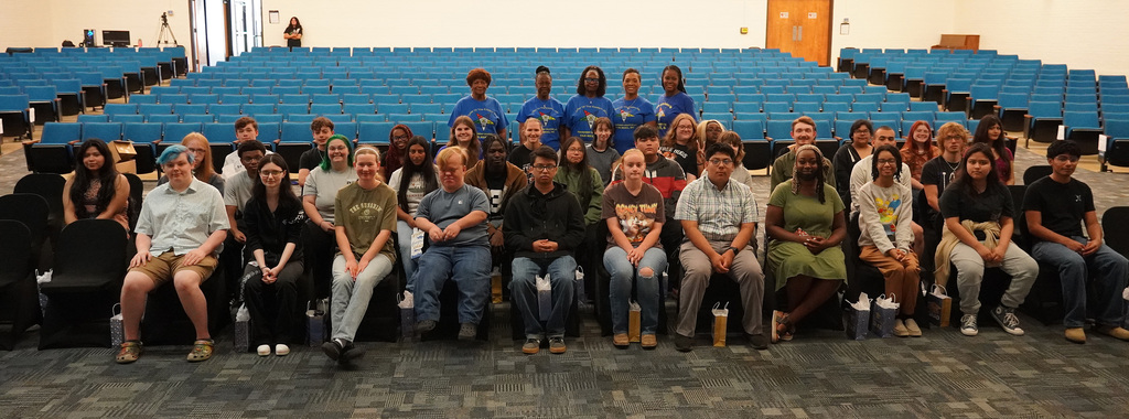 A group of students sits together in an auditorium with 5 adult women standing behind them. 