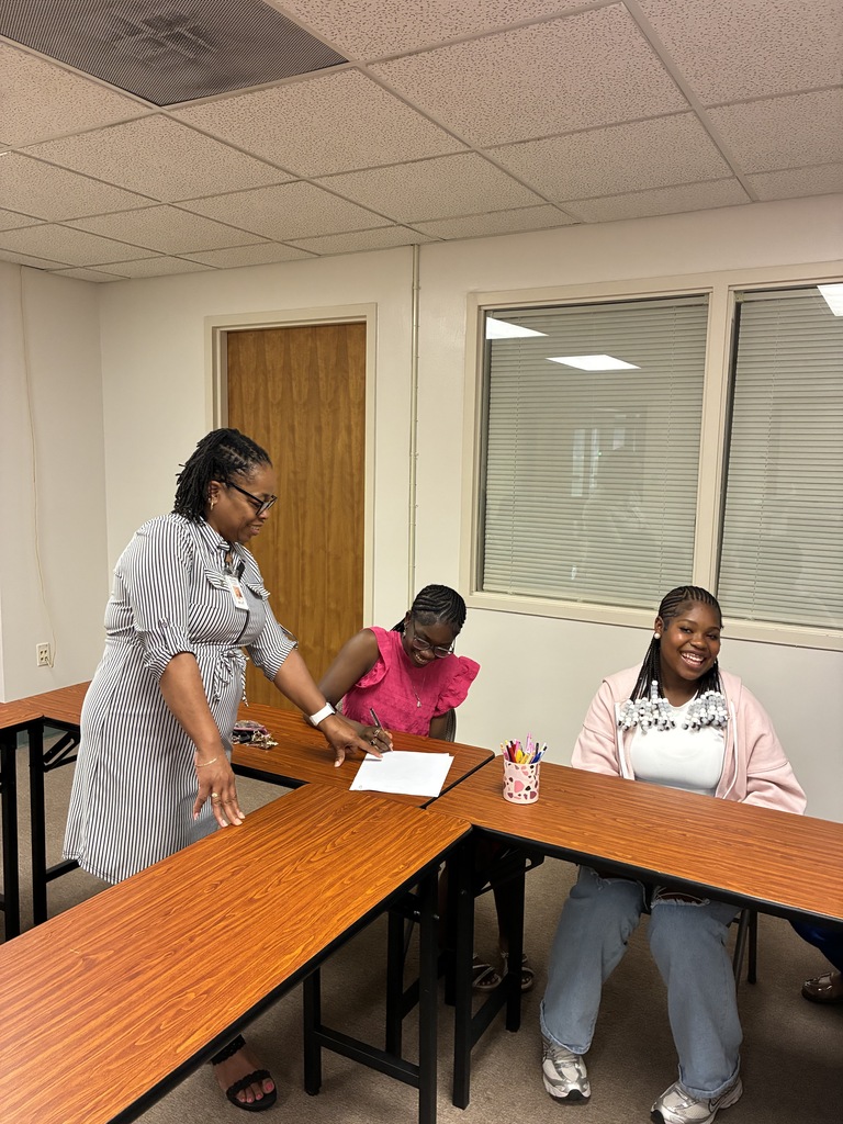 A woman stands beside two smiling girls seated at wooden tables in a classroom or office training space. One girl in a bright pink top is writing on a sheet of paper while the other, wearing a light pink hoodie and jeans, smiles toward the camera. A cup filled with colorful pens sits on the table, and windows with closed blinds line the wall behind them.