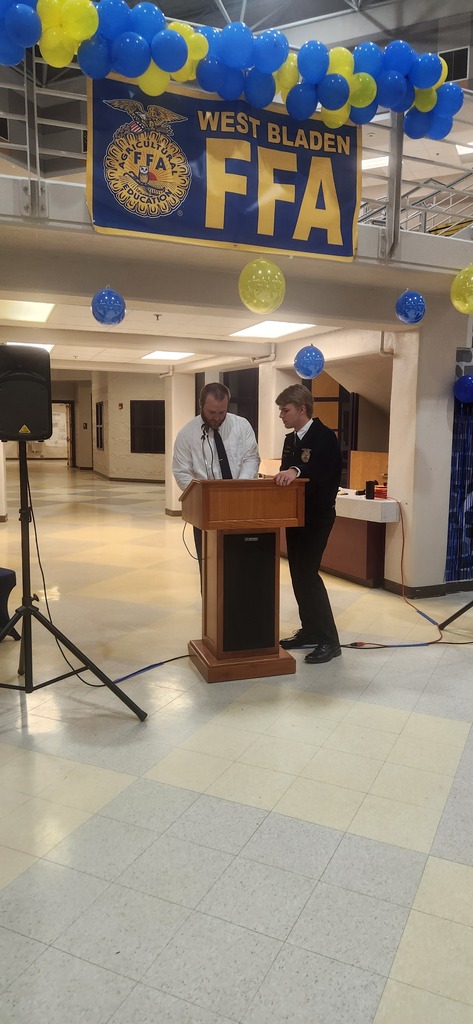 A student officer in an FFA jacket speaking at a podium during the West Bladen FFA banquet, with another student standing nearby on a decorated stage.
