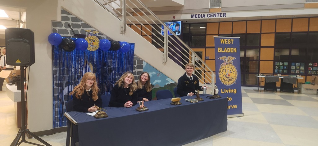 A group of students in blue FFA jackets sitting at a long table decorated with blue table runners, with a staircase and blue and yellow balloon decorations visible in the background