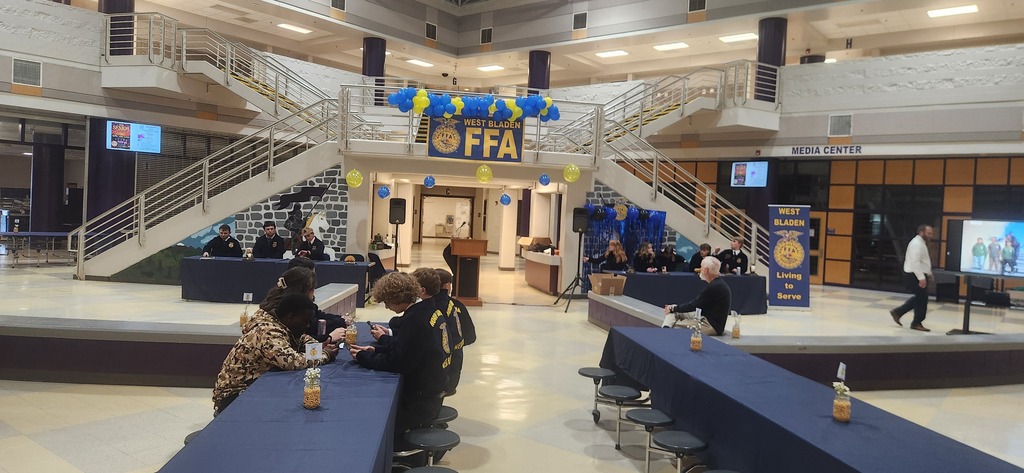 Alt Text: A wide view of a school media center decorated with blue and yellow balloons and a West Bladen FFA sign. Students in official dress are seated at long tables, and parents or guests are gathered in the background