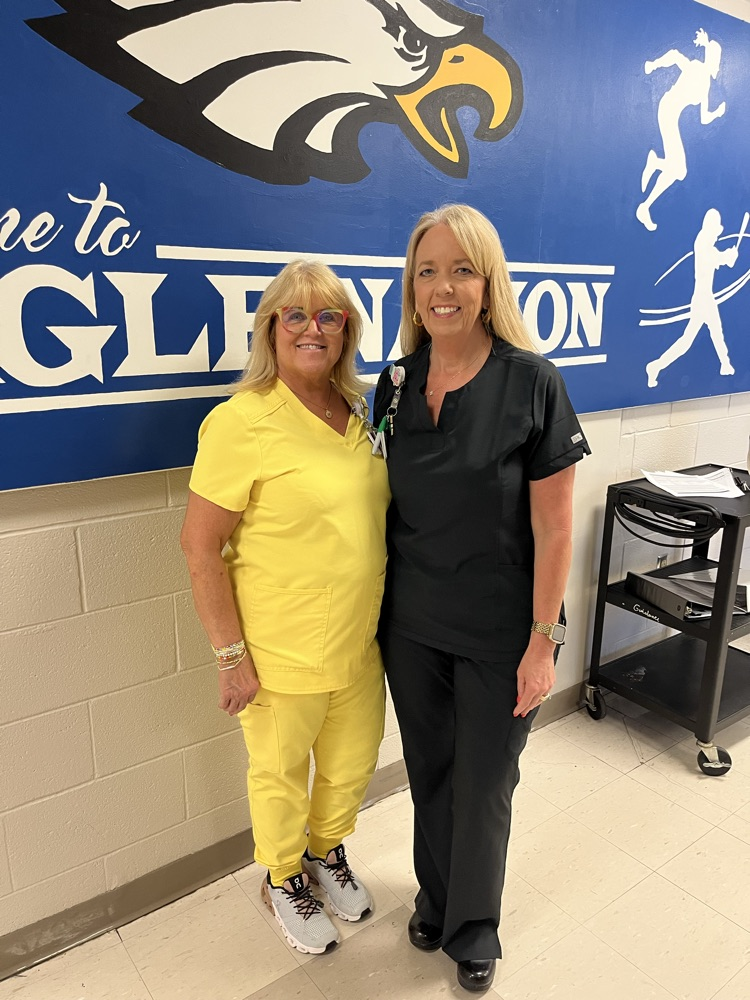 A woman in yellow scrubs stands next to a woman in black scrubs in front of a eagle nation sign