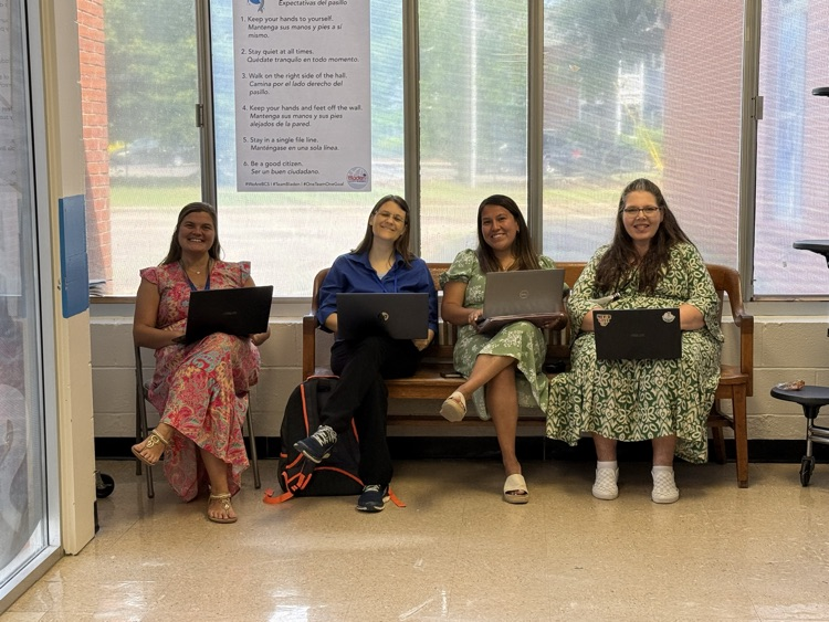 Four first grade teachers sit together in a school hallway with laptops, collaborating and working while classrooms are being used for testing. Large windows and hallway expectation posters are visible in the background.