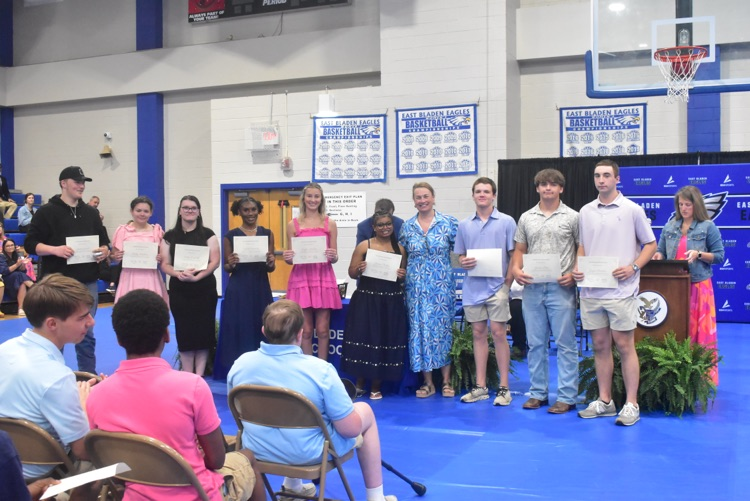 non-student stand with two teachers being present certificate. Students are as professional professionally. There is a blue tarp on the ground and a lady in the back standing in front of a podium.