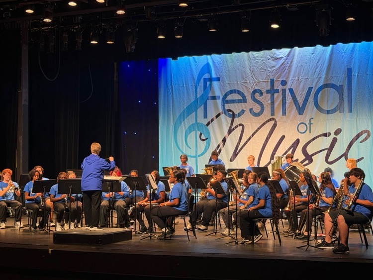 A group of band students performing on stage with their conductor leading in front of them.
