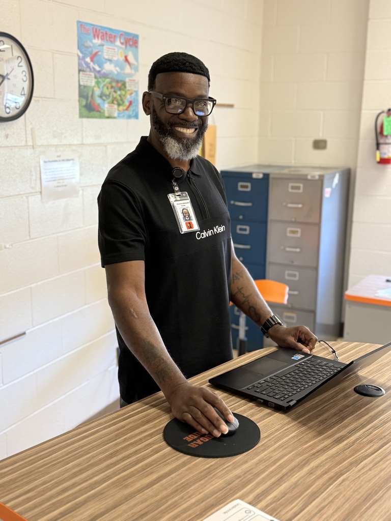 A male staff member smiles while standing at a desk with a laptop in a school classroom setting.