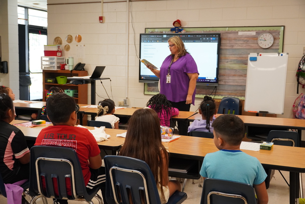 A female teacher in a purple top reads aloud from a book to a group of elementary students seated at desks, with a smartboard displayed at the front of the classroom.
