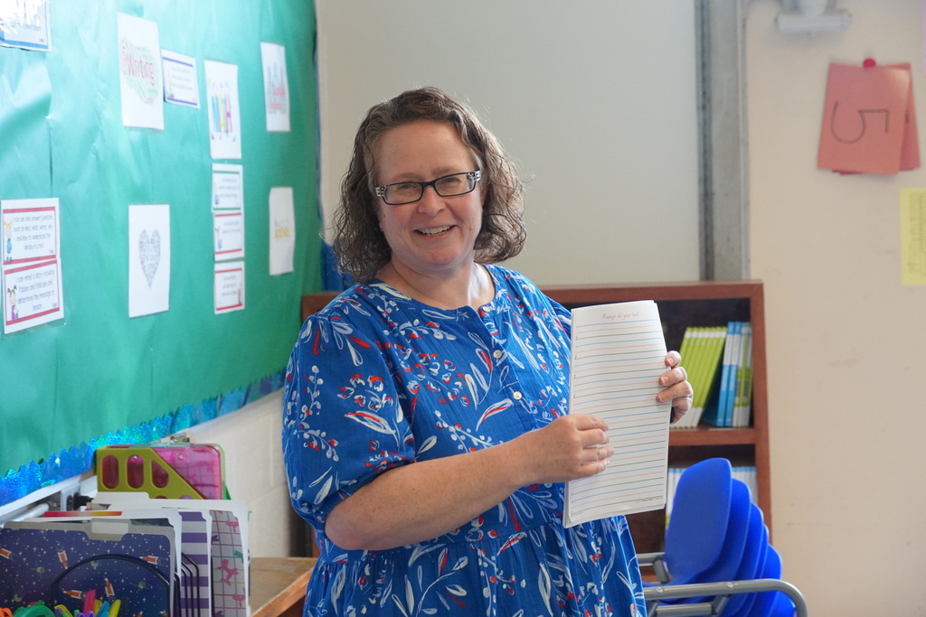A smiling female teacher in a blue floral dress holds up a lined notebook page in a decorated elementary school classroom.