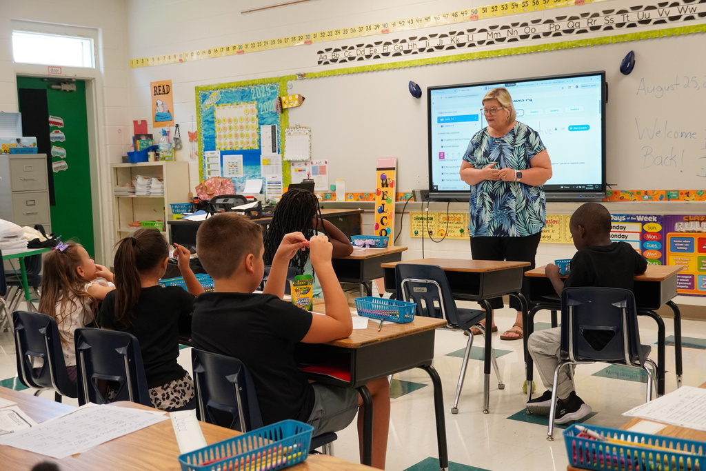  A female teacher stands at the front of a colorful elementary classroom, leading a lesson with students seated at desks and a smartboard displayed behind her.