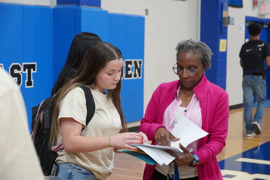 A female teacher is standing with a student reviewing paperwork.