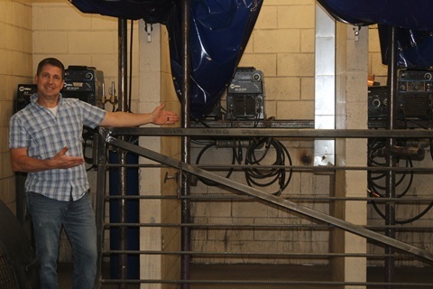 Faculty member Clayton Dowless stands beside a partially completed metal gate in the school’s welding shop, gesturing toward the project. The gate, which includes a steel frame and horizontal metal bars, was constructed with the assistance of students as part of the Class of 2026 senior gift.