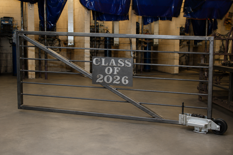 Digital rendering of a metal sliding gate featuring a “Class of 2026” plaque mounted in the center. The gate is constructed from steel tubing and horizontal metal bars and is shown inside a welding and fabrication shop.
