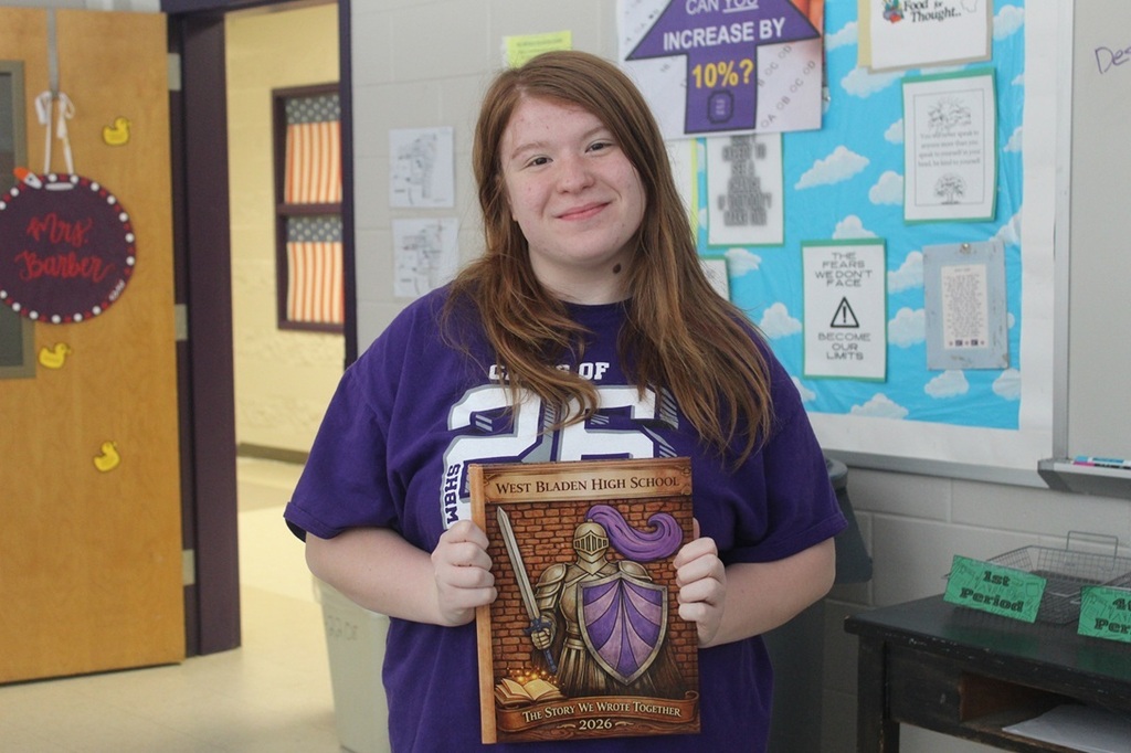 Student stands in a classroom holding a section of the 2026 yearbook cover featuring a knight illustration and shield. The artwork displayed in the cover design was inspired by the student's original artwork.