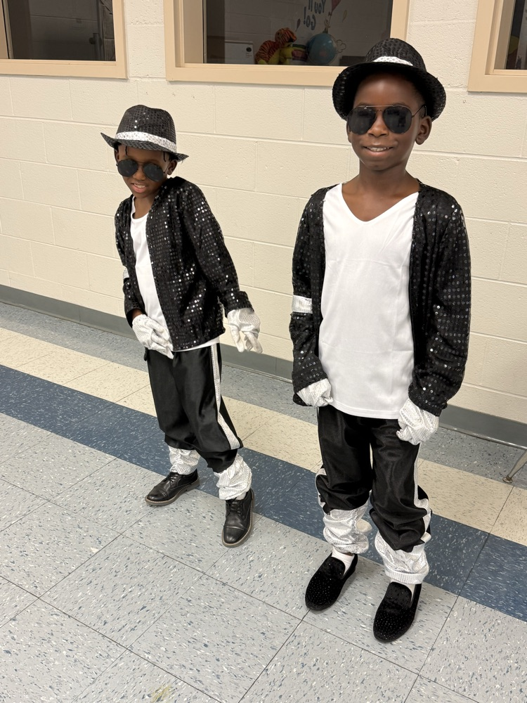Two children wearing Michael Jackson-inspired outfits with black sequined jackets, hats, sunglasses, white gloves, and black pants pose in a school hallway on the last day of school.