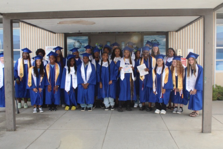 A large group of students in blue cap and gown stand in front of a primary school