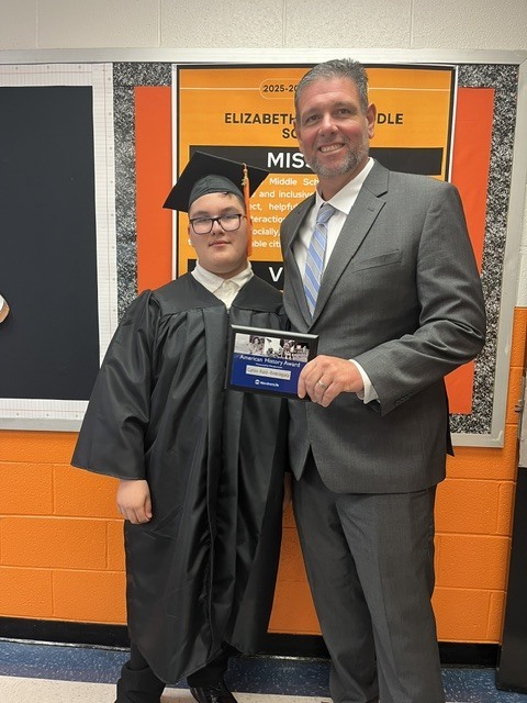 A student wearing a graduation gown and cap stands beside a school administrator while holding a plaque recognizing the American History Award. They are smiling in front of an Elizabethtown Middle School mission statement display featuring the school's orange and black colors.