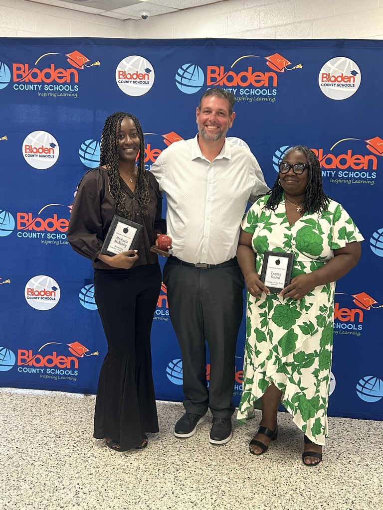 Three educators stand smiling in front of a Bladen County Schools backdrop. Two teachers hold recognition awards for Teacher of the Year and Beginning Teacher of the Year, while the administrator standing between them holds an apple-shaped award. The group is being recognized for their outstanding contributions to education and student success.