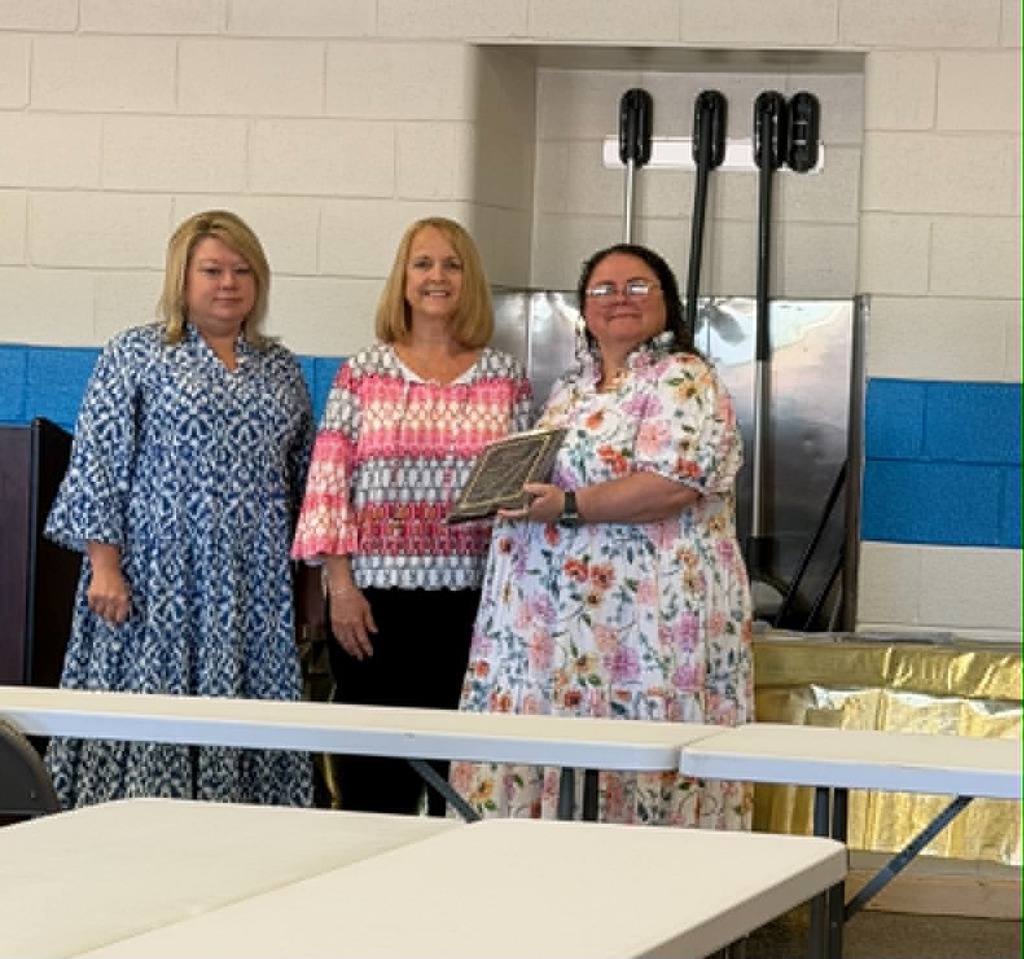 Three staff members stand together during a retirement recognition ceremony in a school cafeteria. One retiree holds a commemorative plaque while colleagues stand beside her. Tables and school signage are visible in the background as the group is honored for their years of service.