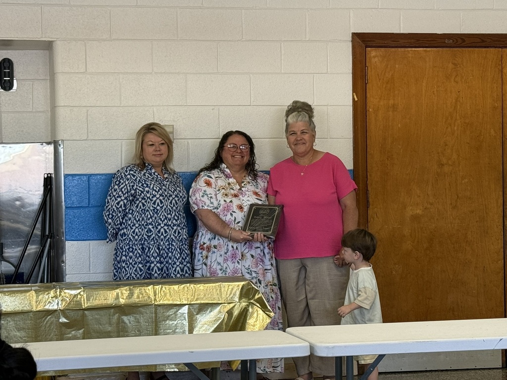 Three staff members stand together during a retirement recognition ceremony in a school cafeteria. One retiree holds a commemorative plaque while colleagues stand beside her. Tables and school signage are visible in the background as the group is honored for their years of service.