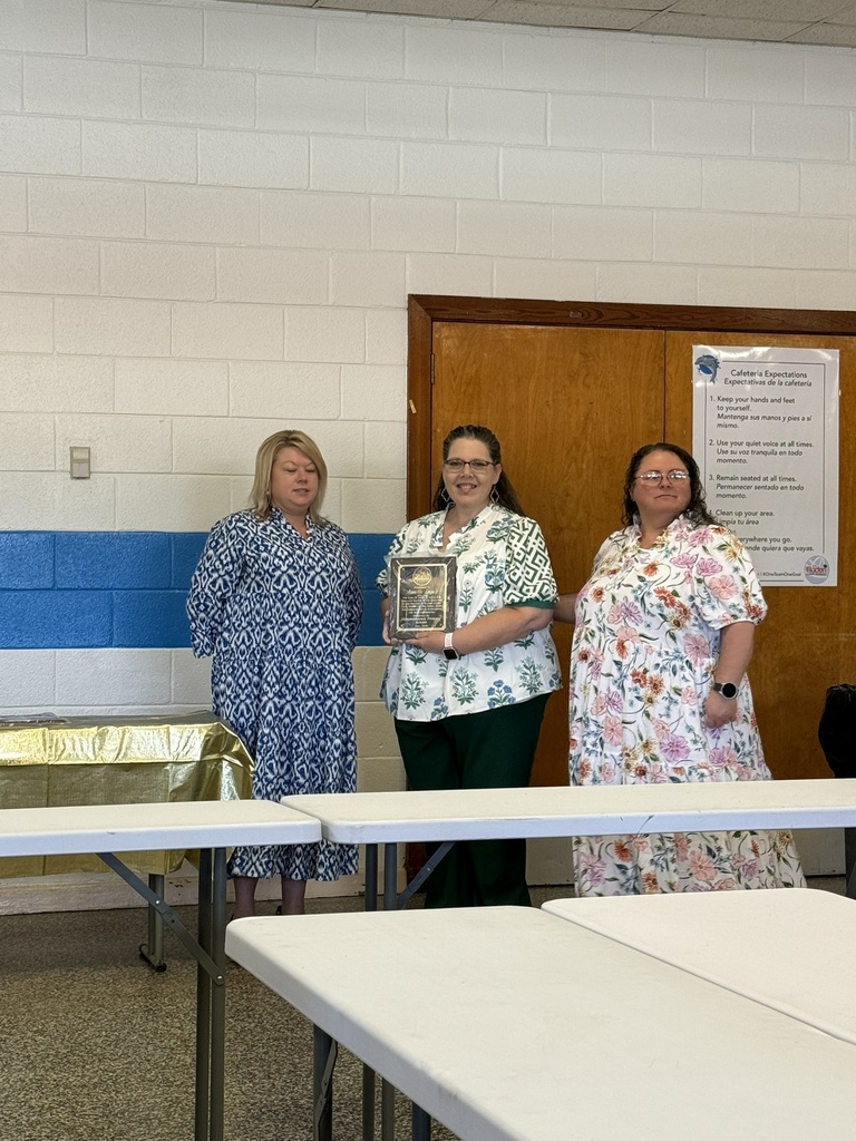 Three staff members stand together during a retirement recognition ceremony in a school cafeteria. One retiree holds a commemorative plaque while colleagues stand beside her. Tables and school signage are visible in the background as the group is honored for their years of service.
