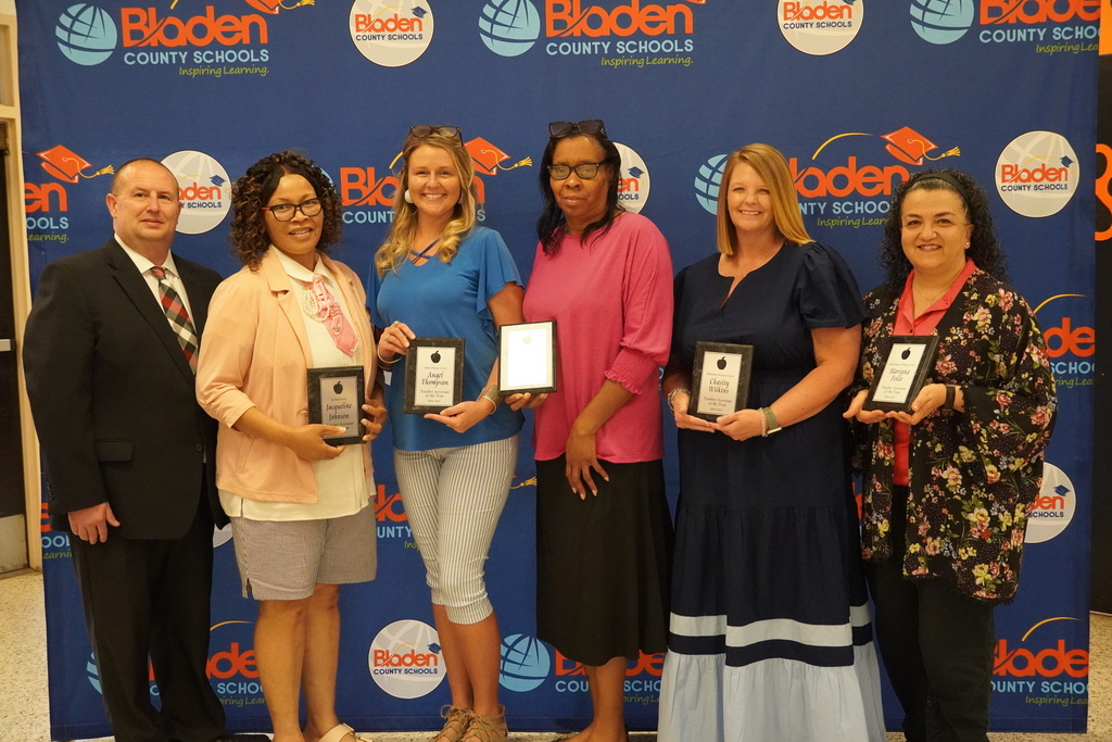 A group of educators stand together smiling and holding plaques in front of a Bladen County Schools backdrop.