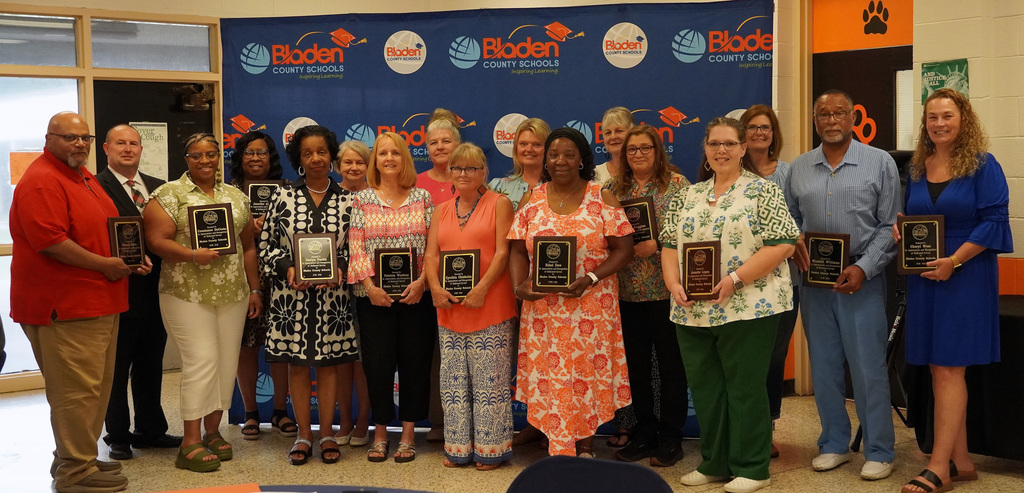 A group of educators stand together smiling, holding plaques in front of a Bladen County Schools backdrop.
