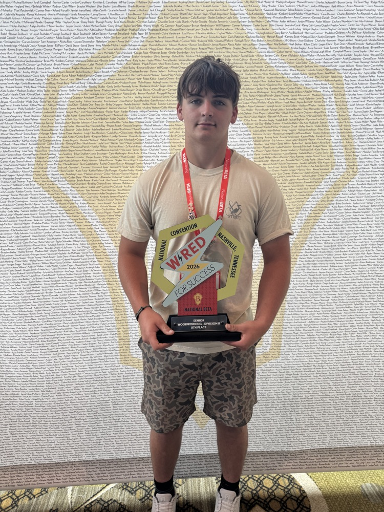 student in camo shorts in front of a Beta backdrop holding a trophy