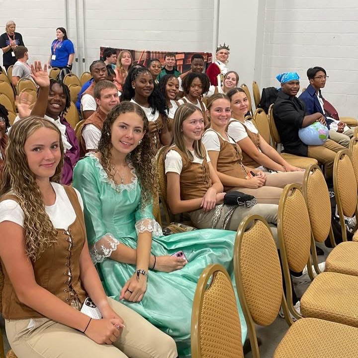 A group of diverse youth sitting in rows of chairs, dressed in Renaissance or historical costumes, including a girl in a teal gown and others in brown vests and tan pants.