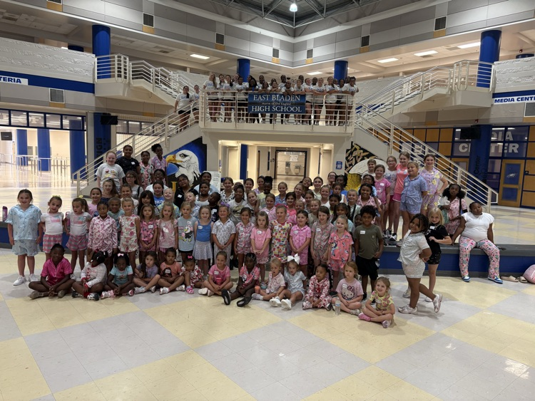 A large group of small children, all girls, but one in pajamas with name tags and a group of cheerleaders standing at the top of stairs posing for a group photo for cheer camp